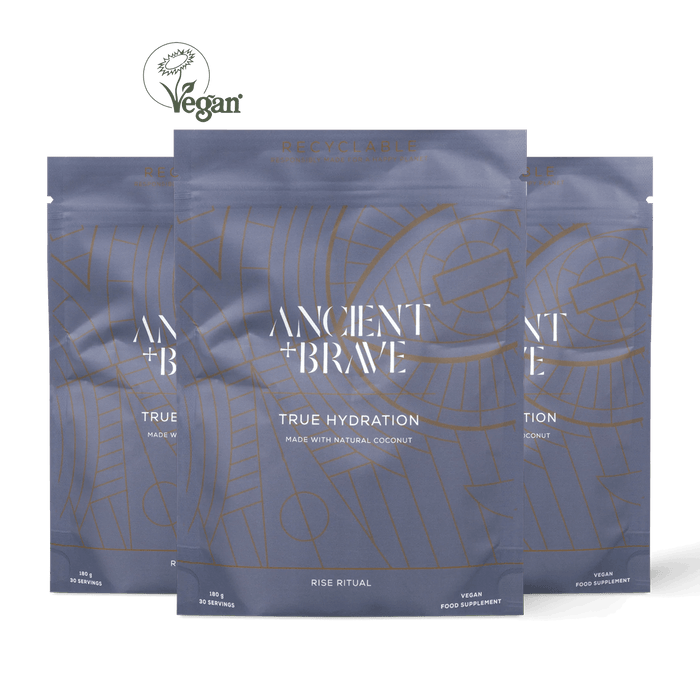 Three stand-up pouches of Ancient + Brave True Hydration food supplement arranged in a fan shape against a solid black background. The packaging is a muted slate blue-gray with white text and gold line-art patterns.